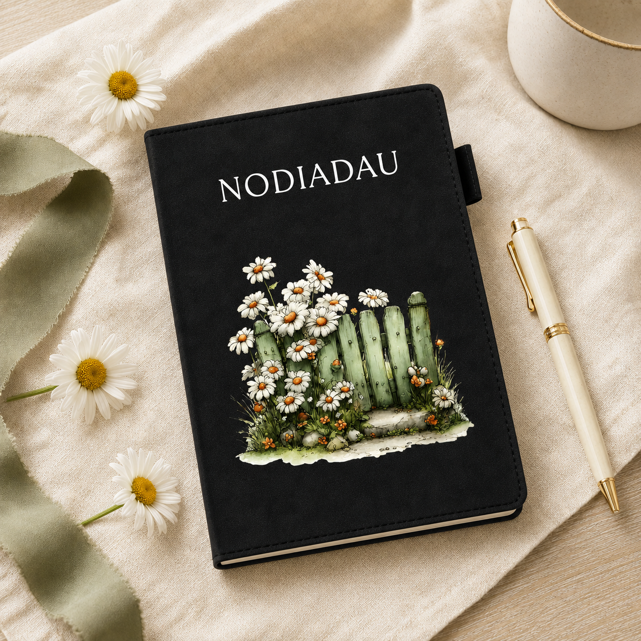 A black hardcover journal with the word 'NODIADAU' printed in white at the top and an illustration of white daisies, green cacti, and small orange flowers on the front. The journal is styled on a beige fabric surface with white daisy flowers, a green ribbon, a cream pen, and part of a white mug visible around it.