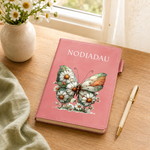 A pink hardcover journal with the name 'NODIADAU' printed in white at the top and a large butterfly made of white daisies with yellow centers on the front cover, styled on a wooden table next to a pen and a vase of daisies.