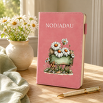 A pink hardcover notebook with the word 'NODIADAU' printed on the cover, decorated with a floral illustration of white daisies and a green sign. The notebook stands on a wooden table next to a vase of white flowers and a gold pen.