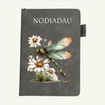 A dark gray faux-leather notebook or journal cover featuring a printed illustration of a green dragonfly resting on white daisies. The word 'NODIADAU' is printed in white serif letters at the top.
