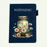 A dark navy blue journal or notebook cover featuring the word 'NODIADAU' in white serif text at the top and an illustration of a glass jar filled with glowing fireflies and white daisies on the front. A small tab is visible on the right side.