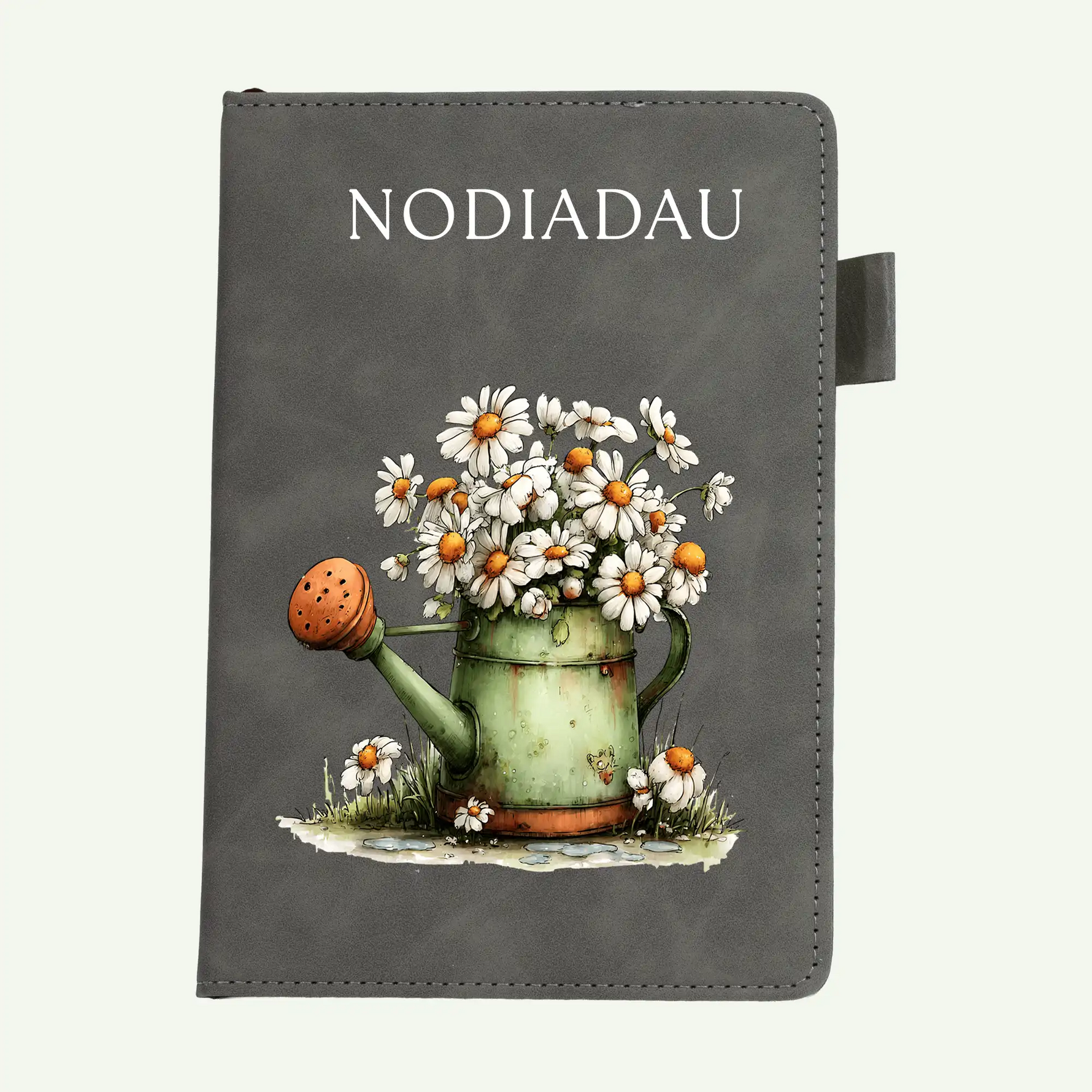 A dark gray notebook or journal cover featuring the word 'NODIADAU' in white serif text at the top and an illustration of a green watering can filled with white daisies and small flowers. A black tab extends from the right side.