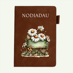 A brown faux-leather notebook cover with the word 'NOTEBOOK' printed at the top and an illustration of white daisies with orange centers, a rustic wooden sign, and small wildflowers on the front, shown against a light off-white background.